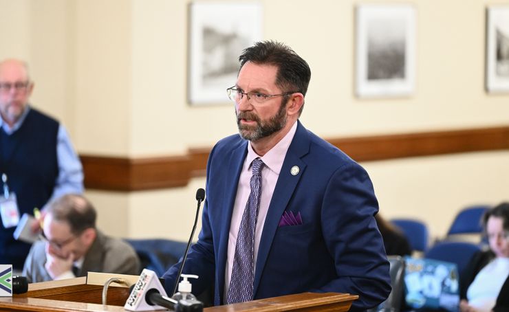 Marshall, a man with light-toned skin, wears a blue suit jacket and glasses as he stands behind a podium.  On person stands behind him and three are sitting.  