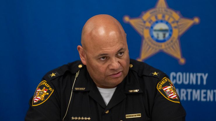 Sheriff Harold Pretel, a man with medium-toned skin, wearing a sheriff's uniform, sits in front of a blue background with a sheriff's star printed on it.  